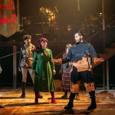 Actors in post-apocalyptic costumes on stage in Urinetown, with the band visible on a raised level in the background